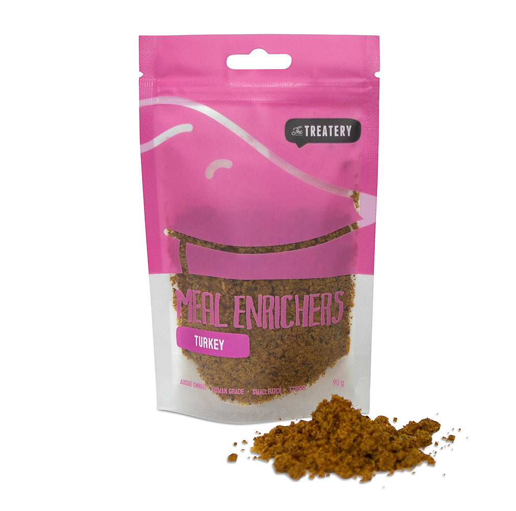 A pink package of 'The Treatery' brand Turkey Meal Enrichers for dogs. The package shows a clear window revealing the brown powder product.